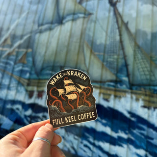 Sticker with 'Wake the Kraken Full Keel Coffee' design held in front of a ship painting.