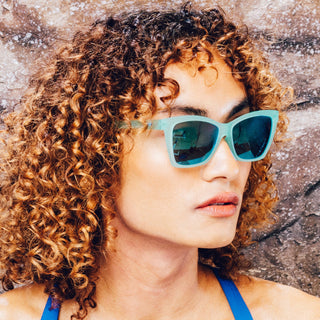 Woman wearing blue goodr sunglasses and a blue top against a stone wall background