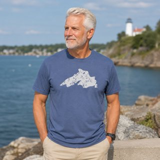 Man wearing a blue t-shirt with a white graphic design, standing by a body of water with a lighthouse in the background. AdventureUs Lake Superior Vintage Print T Shirt - Lifestyle