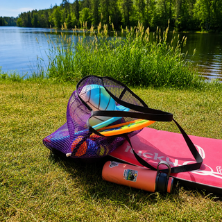 Mesh tote filled with volleyball, Frisbee, water bottle and towel on a grassy area near a lake with trees in the background. Sand Island mesh beach tote made in Washburn Wisconsin for beach days, sports gear, towels, and lake adventures.