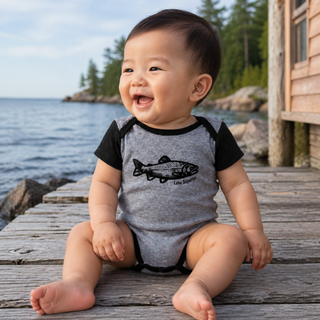 Baby sitting on a wooden dock by the water wearing a gray and black onesie with a Lake Superior fish design.