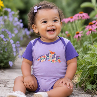 Child wearing a purple onesie with floral design sitting in a garden.