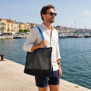 Man holding a black mesh bag by a waterfront with buildings and boats in the background