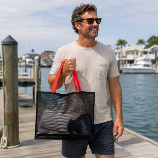Man holding a black mesh tote bag with red handles on a dock by the water.